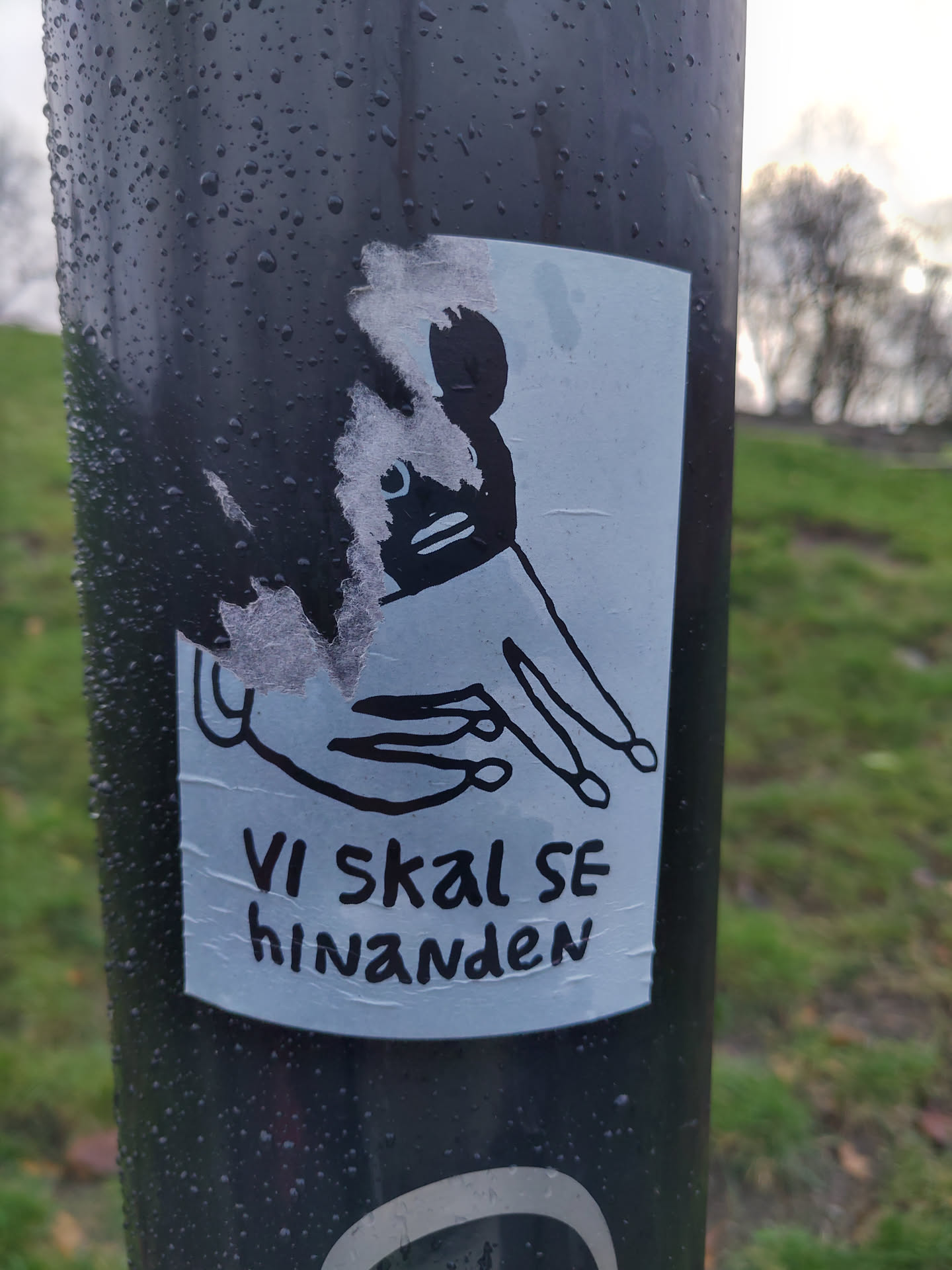 A sticker featuring a simple line drawing of a creature with a hand reaching out. The text below the drawing says "Vi skal se hinanden", which is Danish for "We shall see each other". The sticker is affixed to a dark pole outdoors.