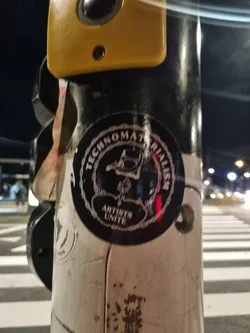 A black and white circular sticker with the text "Technomaterialism" and "Artists Unite" surrounding a central design. The central design appears to be an abstract symbol or logo.