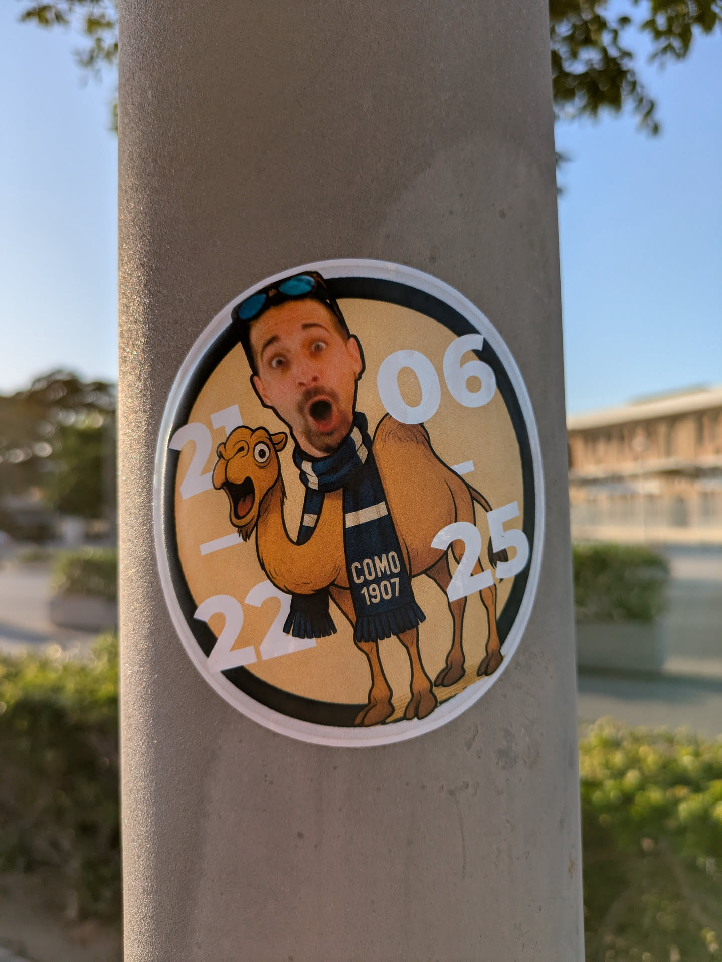 A round sticker is affixed to a gray pole, featuring a cartoon-like image of a man with a surprised expression riding a camel. The camel is wearing a scarf with "COMO 1907" printed on it. The background of the sticker is beige, with the numbers "21", "22", "06", and "25" encircling the image. The sticker has a white border, creating a contrast against the pole.