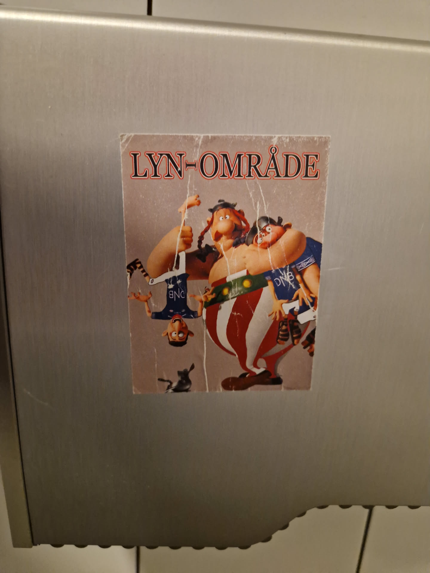 A sticker depicting a humorous cartoon scene of vikings. The text "LYN-OMRÅDE" is visible at the top.  The style is reminiscent of claymation or stop-motion animation. The sticker appears to be affixed to a metallic surface, possibly in a public restroom.