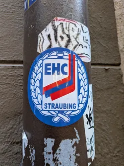 An oval-shaped sticker featuring the emblem of EHC Straubing against a solid blue background. A white laurel wreath borders a central shield-shaped badge. Inside the shield, the bold blue letters 'EHC' sit at the top, followed by diagonal hockey stick motifs in red and blue, and the word 'STRAUBING' printed in blue along the bottom.