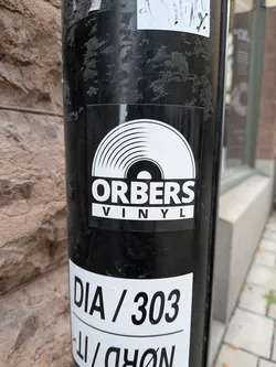 A square, glossy sticker with a black background affixed to a cylindrical black post. At the top of the sticker graphic is a stylized white illustration representing the upper half of a vinyl record with concentric groove lines. Beneath the record illustration is the word 'ORBERS' printed in bold, white uppercase lettering, underlined by a white rectangular banner containing the word 'VINYL' in spaced-out black capital letters.