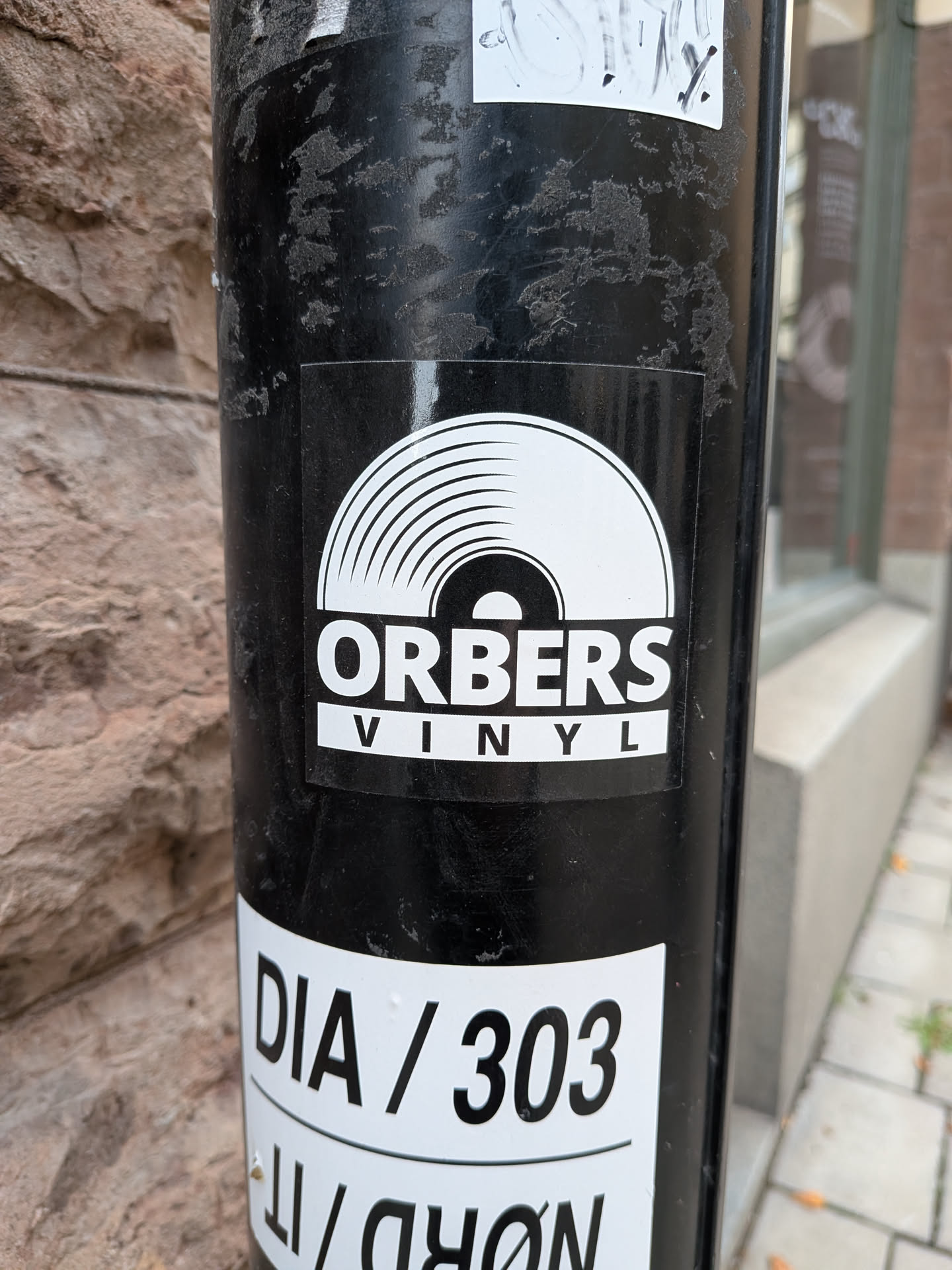 A square, glossy sticker with a black background affixed to a cylindrical black post. At the top of the sticker graphic is a stylized white illustration representing the upper half of a vinyl record with concentric groove lines. Beneath the record illustration is the word 'ORBERS' printed in bold, white uppercase lettering, underlined by a white rectangular banner containing the word 'VINYL' in spaced-out black capital letters.
