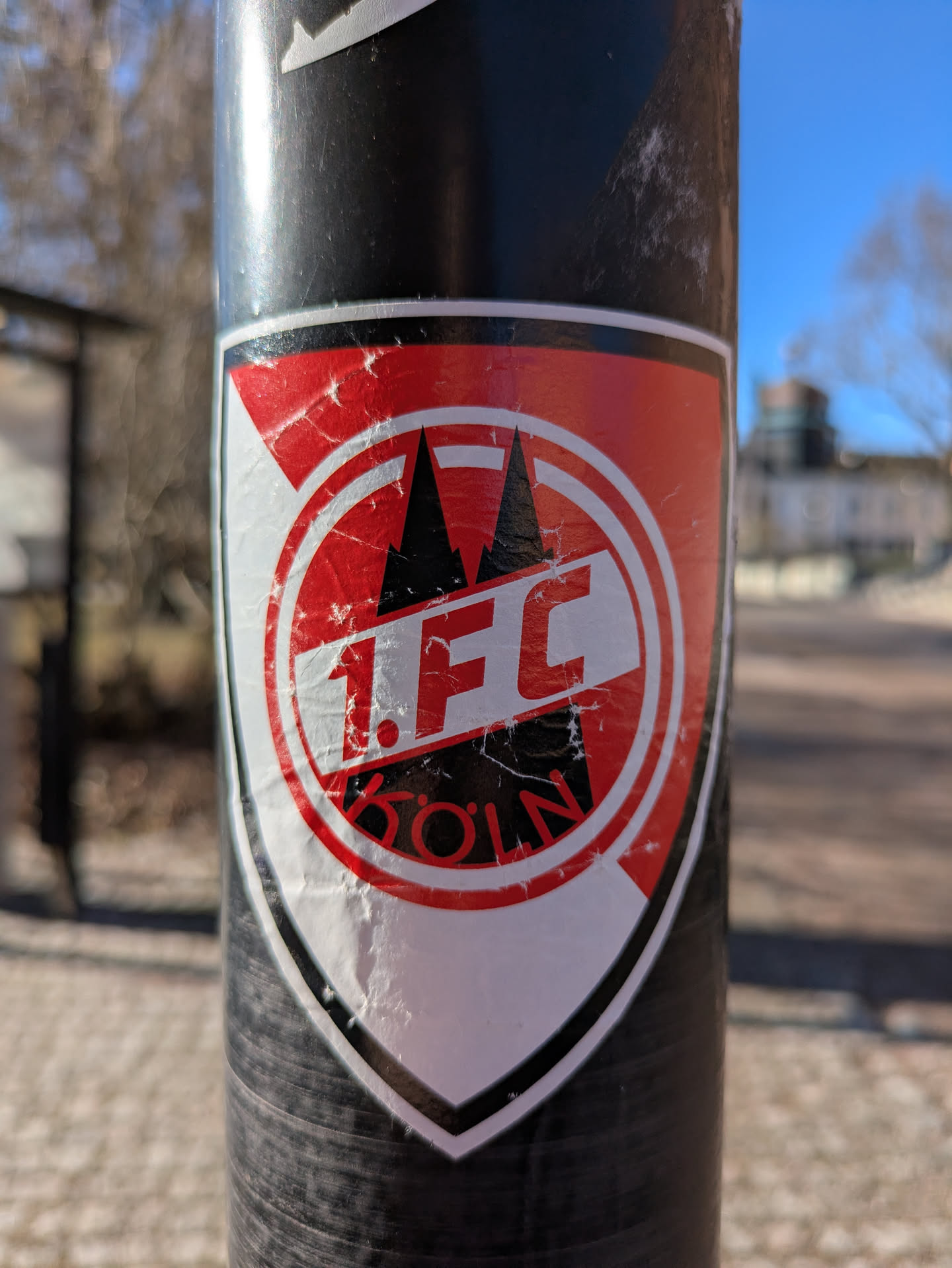 A worn sticker of the 1. FC Köln logo affixed to a black pole. The logo is red and white with the Cologne Cathedral depicted above the text "1. FC KÖLN".