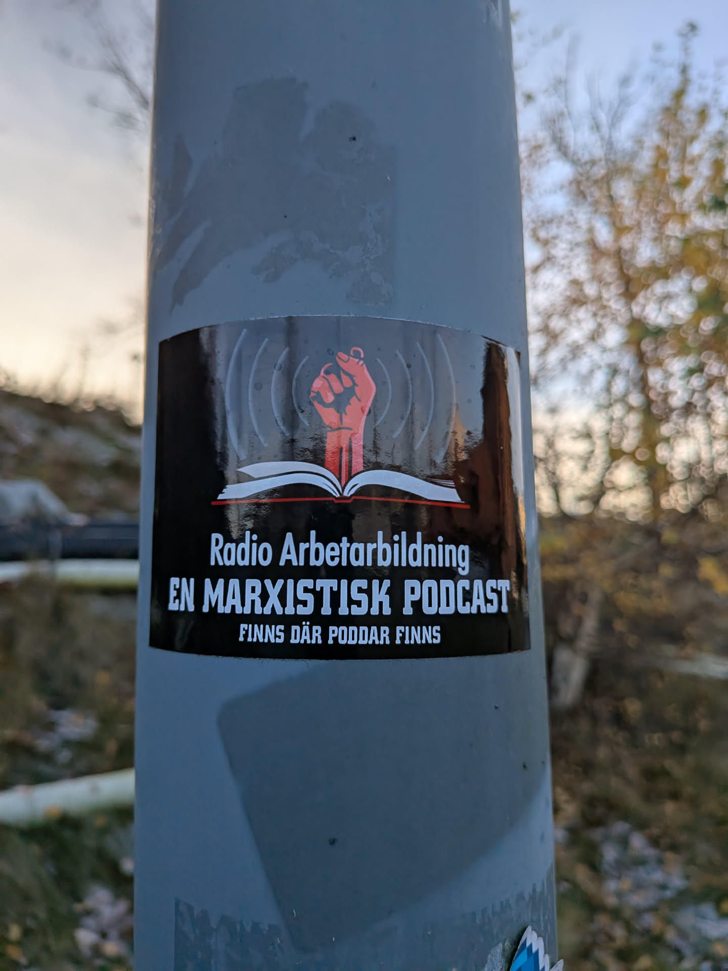 The sticker is rectangular with a black background. At the top, there are stylized grey soundwaves emanating from the center. Below the soundwaves, a red raised fist is depicted emerging from an open book. The book has white pages and a red spine. The text on the sticker is in white, sans-serif font. The top line reads 'Radio Arbetarbildning'. The second line reads 'EN MARXISTISK PODCAST'. The third line reads 'FINNS DÄR PODDAR FINNS'.