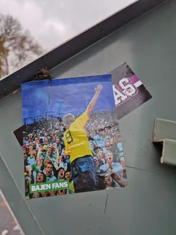 A photo sticker of a football player raising his arm in celebration, surrounded by enthusiastic fans. The text 'BAJEN FANS' is printed at the bottom. The background is a blurry stadium.