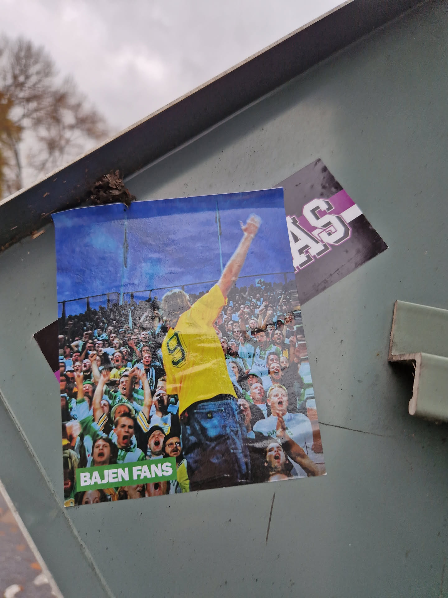 A photo sticker of a football player raising his arm in celebration, surrounded by enthusiastic fans. The text 'BAJEN FANS' is printed at the bottom. The background is a blurry stadium.