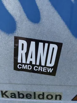 A black sticker with white uppercase lettering reading "RAND CMD CREW". The sticker is affixed to a light gray surface with blue spray paint in the background.