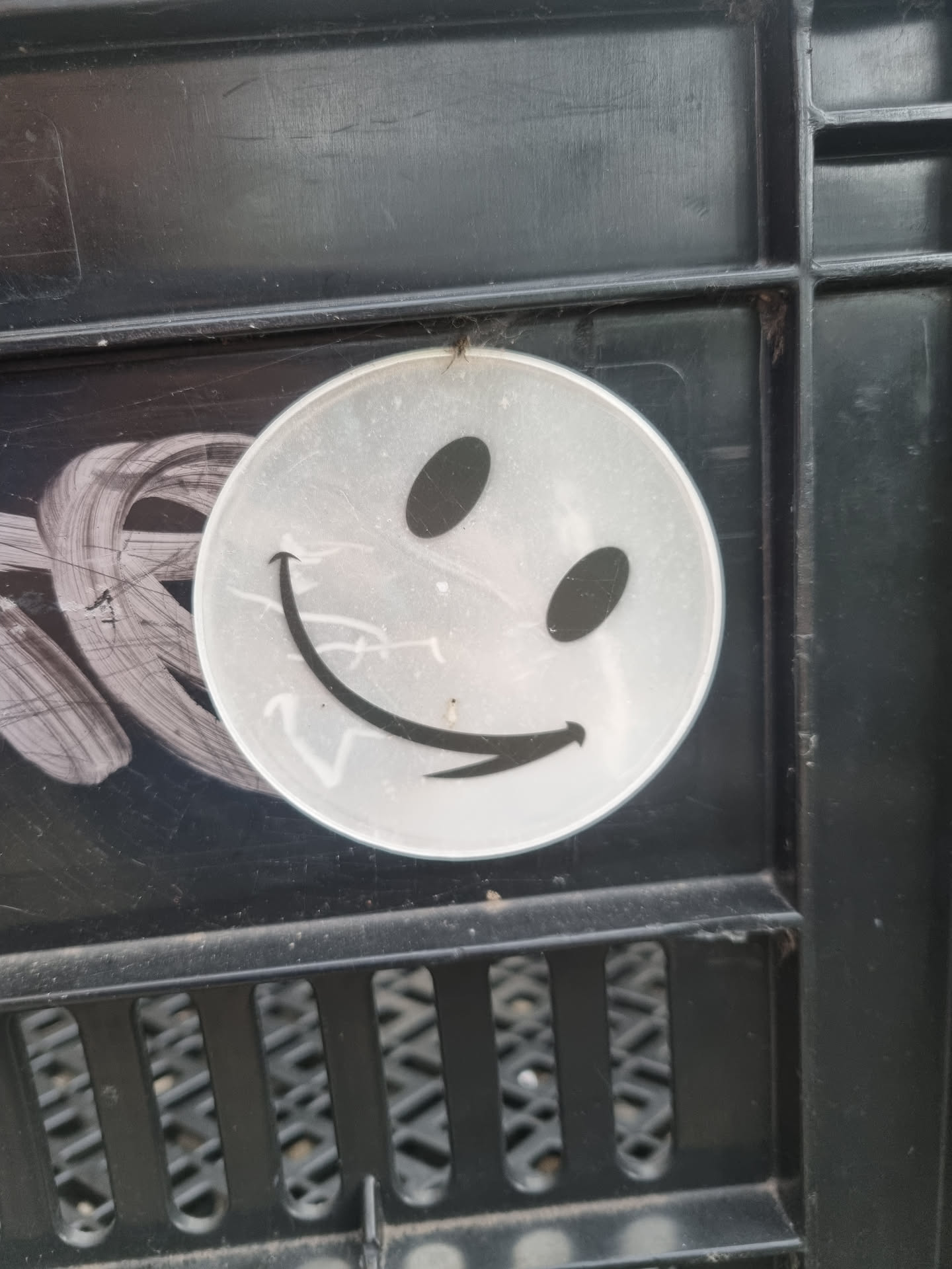 A simple, round sticker with a slightly mischievous smiley face design.  The sticker is white or light gray with black features and appears to be applied to a dark gray or black plastic surface.