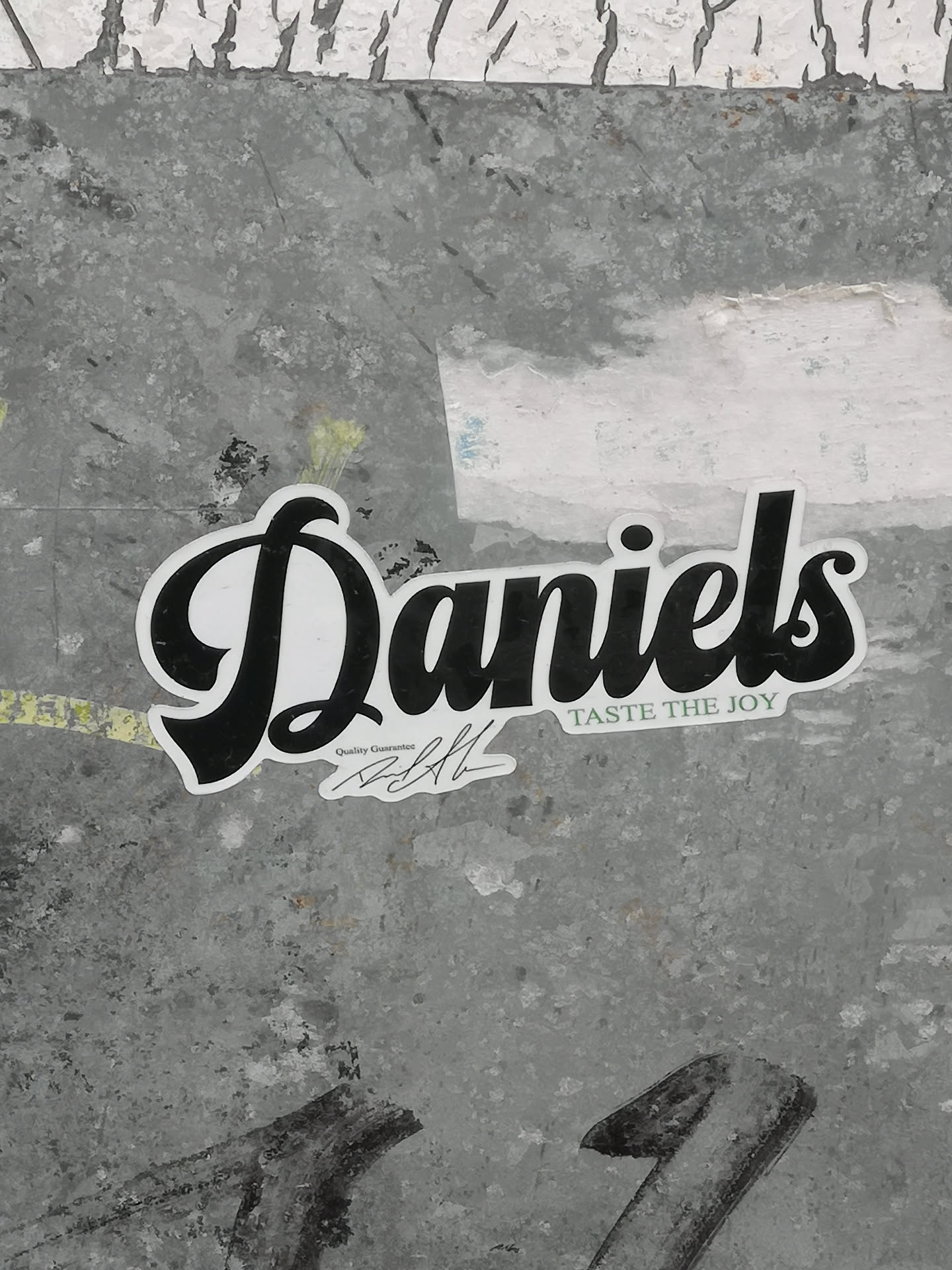 The sticker prominently displays the word 'Daniels' in a bold, flowing black script font with a clean white fill and a thin black outline. Below 'Daniels' and slightly to the right, the slogan 'TASTE THE JOY' is written in a smaller, sans-serif font. Further below and to the left, in an even smaller font, are the words 'Quality Guarantee' followed by a stylized black signature. The sticker has a distinct white border around all its elements, making it stand out against the textured, mottled gray background it's adhered to.