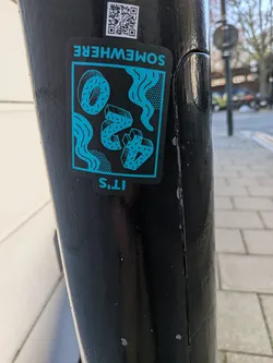 A rectangular sticker with rounded corners, featuring a striking two-tone design in black and a bright teal/cyan. The background is black, while all illustrative elements are rendered in teal. The central image depicts a surreal composition of objects: a whole donut with sprinkles in the upper left, two stylized or broken donut-like shapes with sprinkles in the middle, and two geometric, crystal-like forms with dotted textures on the right. Wavy lines and dotted patterns flow throughout the background, creating a dynamic, liquid-like effect. Above the main graphic, the word "SOMEWHERE" is written upside down in teal, and below it, "IT'S" is also written upside down in teal, making the full phrase "IT'S SOMEWHERE" when viewed correctly.