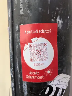 A red Sciencicast sticker is affixed to a black pole. The sticker features the question 'A corto di scienza?' which translates to 'Short on science?'. A QR code is prominently displayed, along with the Instagram handle @SCICAST. The sticker also includes the text 'Ascolta Scientificast!' meaning 'Listen to Scientificast!'. A cartoon atom symbol is included in the design. There is also a torn piece of a sticker beneath it
