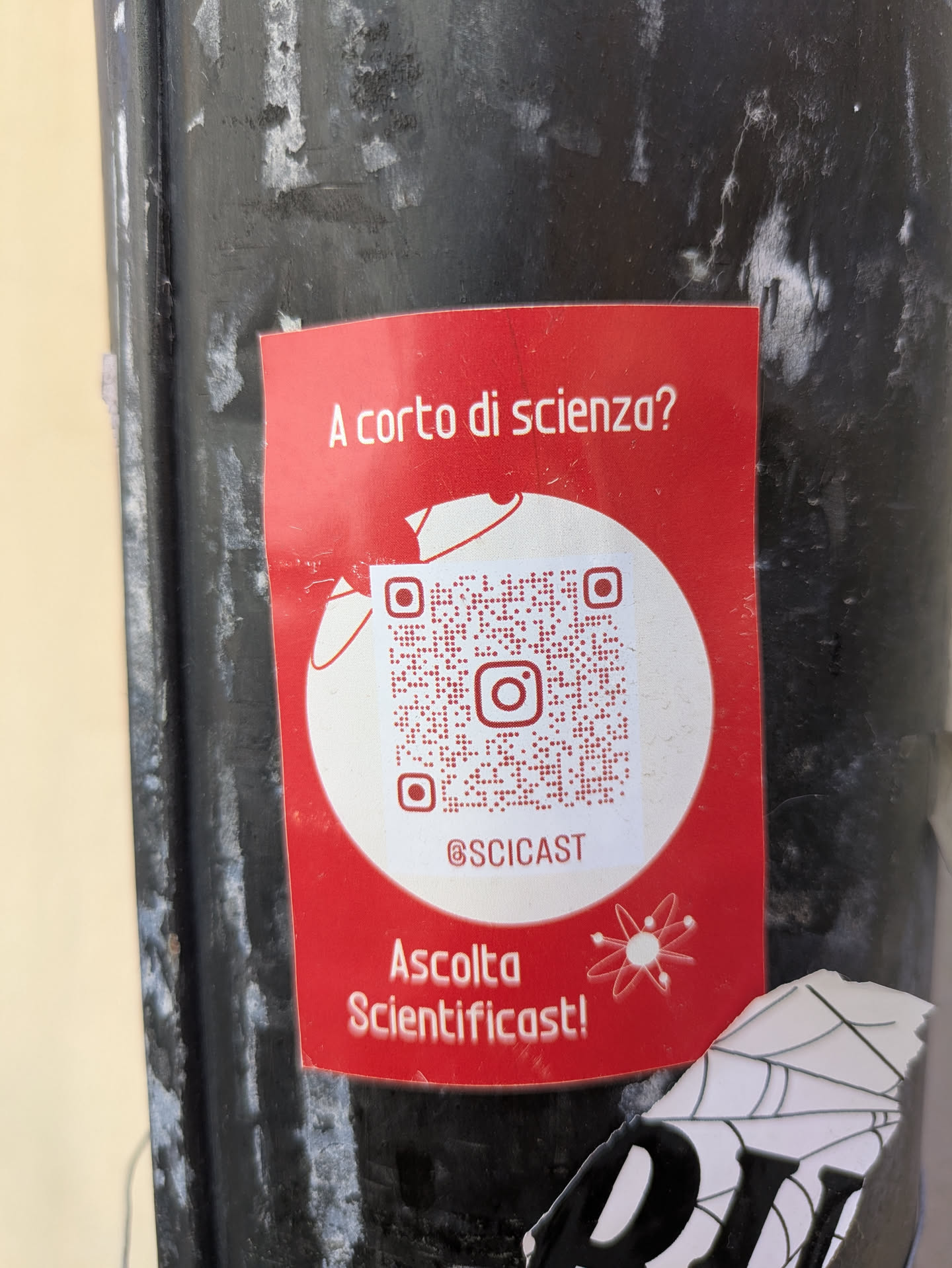 A red Sciencicast sticker is affixed to a black pole. The sticker features the question 'A corto di scienza?' which translates to 'Short on science?'. A QR code is prominently displayed, along with the Instagram handle @SCICAST. The sticker also includes the text 'Ascolta Scientificast!' meaning 'Listen to Scientificast!'. A cartoon atom symbol is included in the design. There is also a torn piece of a sticker beneath it