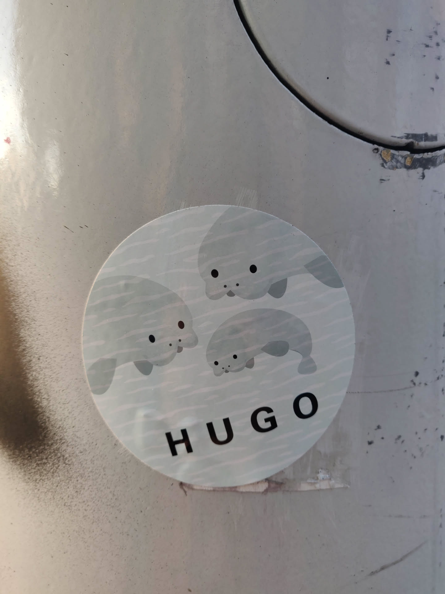 A round sticker featuring an illustration of three adorable cartoon manatees swimming. The word "HUGO" is printed underneath the manatees. The sticker appears to be affixed to a light gray surface.