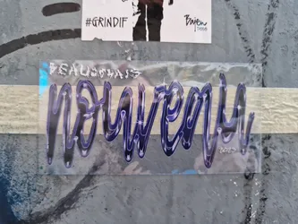 A transparent sticker with the word "Nouveau" written in a stylized, dark blue and purple font. The year 2023 is subtly included at the bottom right.  The sticker appears to be hand-drawn and has a slightly glittery or cosmic effect in its design. It is affixed to a gray, textured surface.