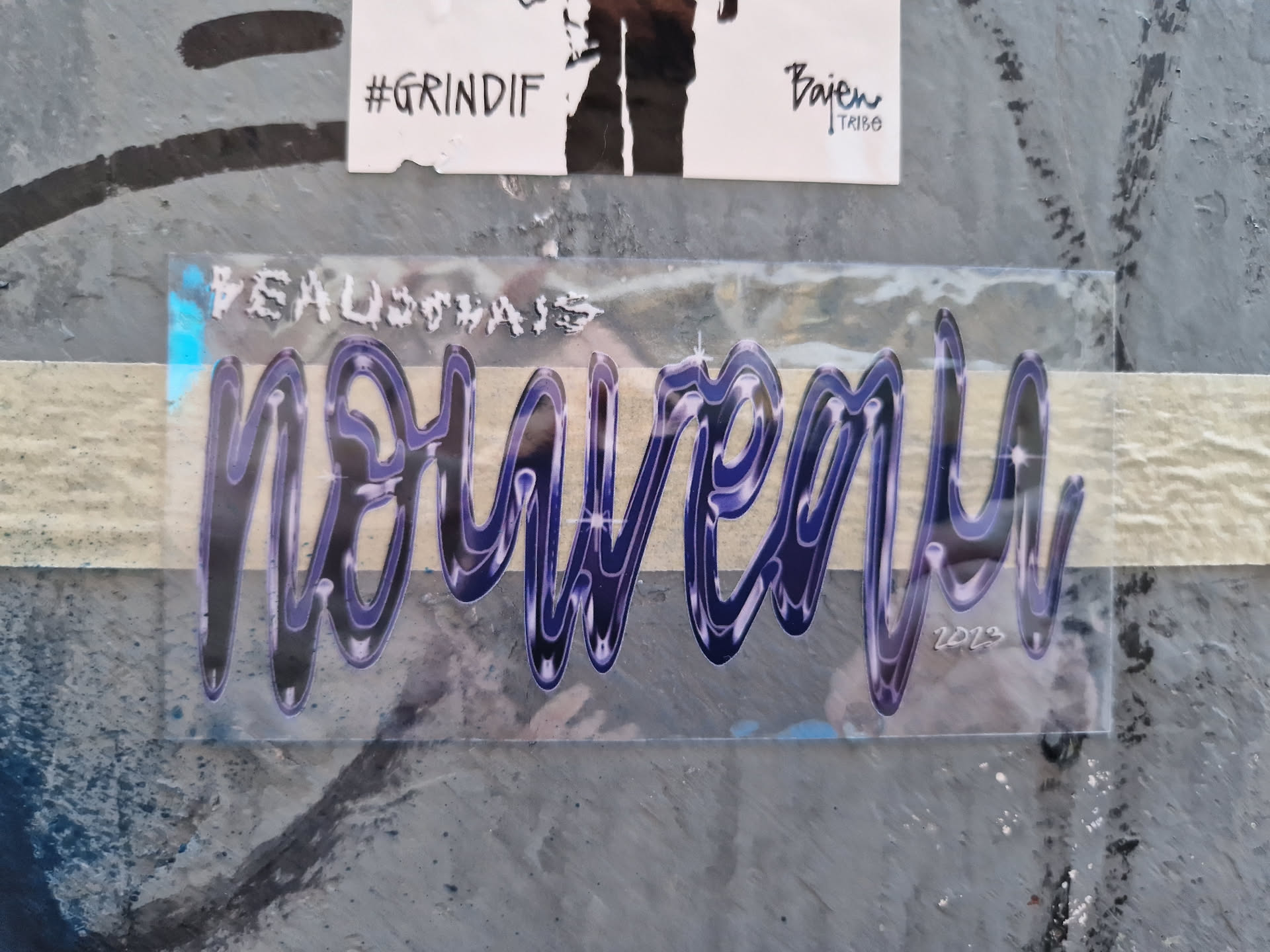 A transparent sticker with the word "Nouveau" written in a stylized, dark blue and purple font. The year 2023 is subtly included at the bottom right.  The sticker appears to be hand-drawn and has a slightly glittery or cosmic effect in its design. It is affixed to a gray, textured surface.