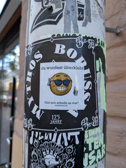 Street sticker A square white sticker placed directly over another sticker on a lamppost or pole. It features a 3D-rendered yellow emoji wearing dark sunglasses, flashing a wide toothy grin while holding up a thumbs-up gesture and touching its shades. Text above the emoji reads 'du wurdest überklebt' and below it reads 'fühlt sich scheiße an was?' followed by the website 'stickerknax.de'.