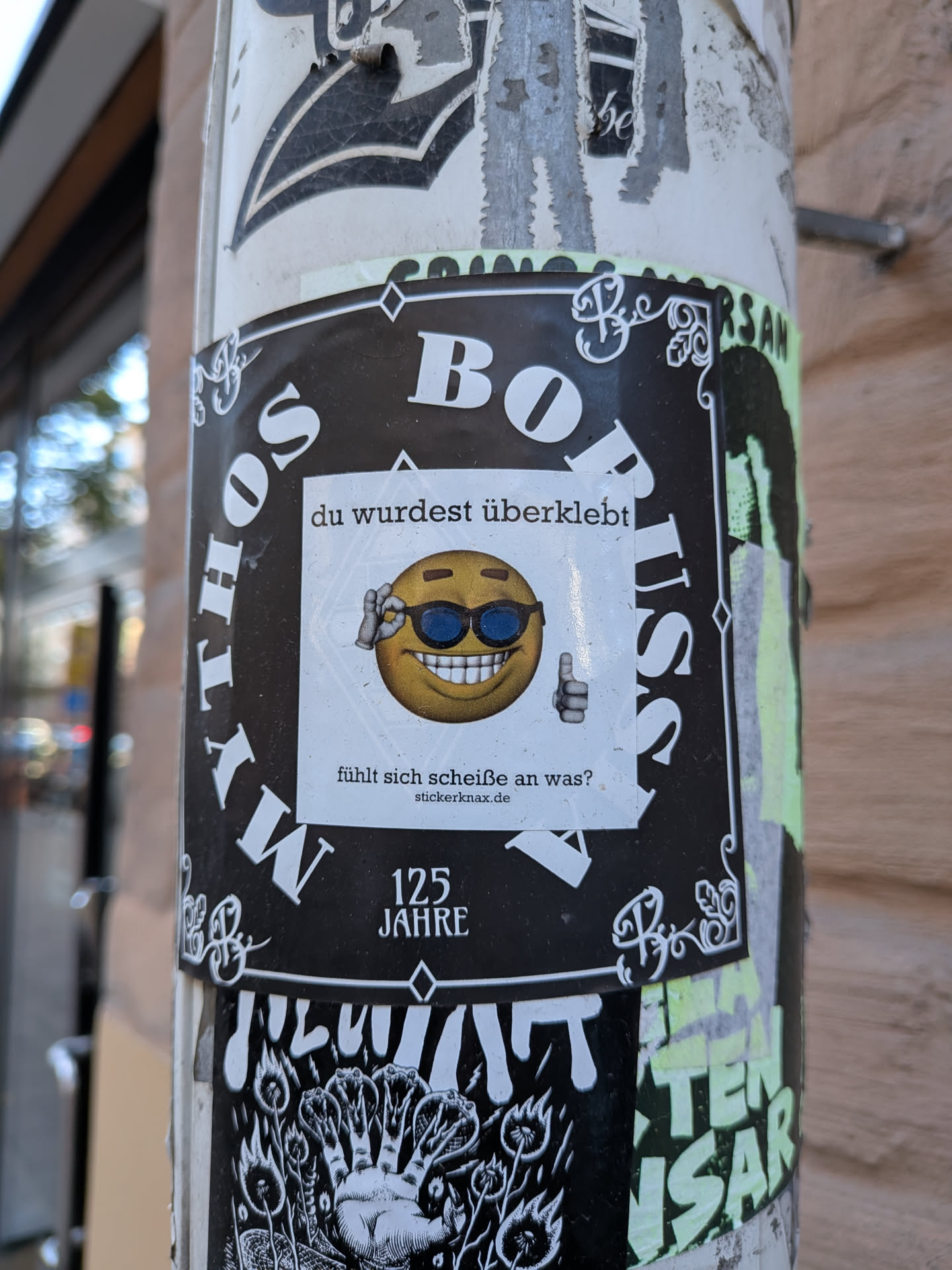 A square white sticker placed directly over another sticker on a lamppost or pole. It features a 3D-rendered yellow emoji wearing dark sunglasses, flashing a wide toothy grin while holding up a thumbs-up gesture and touching its shades. Text above the emoji reads 'du wurdest überklebt' and below it reads 'fühlt sich scheiße an was?' followed by the website 'stickerknax.de'.