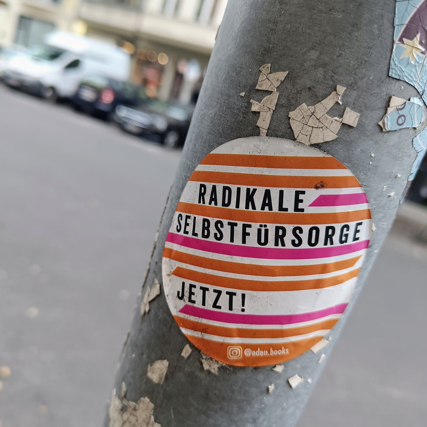 A round sticker with orange and pink stripes. The text on the sticker says "Radikal Selbstfürsorge Jetzt!" which translates to "Radical Self-Care Now!" in German.  There is also an Instagram handle on the bottom of the sticker.