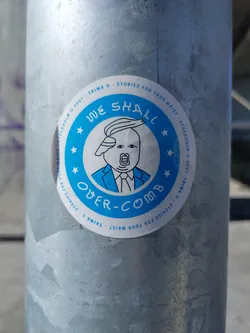 A round blue sticker with a cartoon illustration of Donald Trump in a suit and the text "We Shall Over-Comb". The sticker also features the text "Stories for your wrist" and the brand "Triwa".