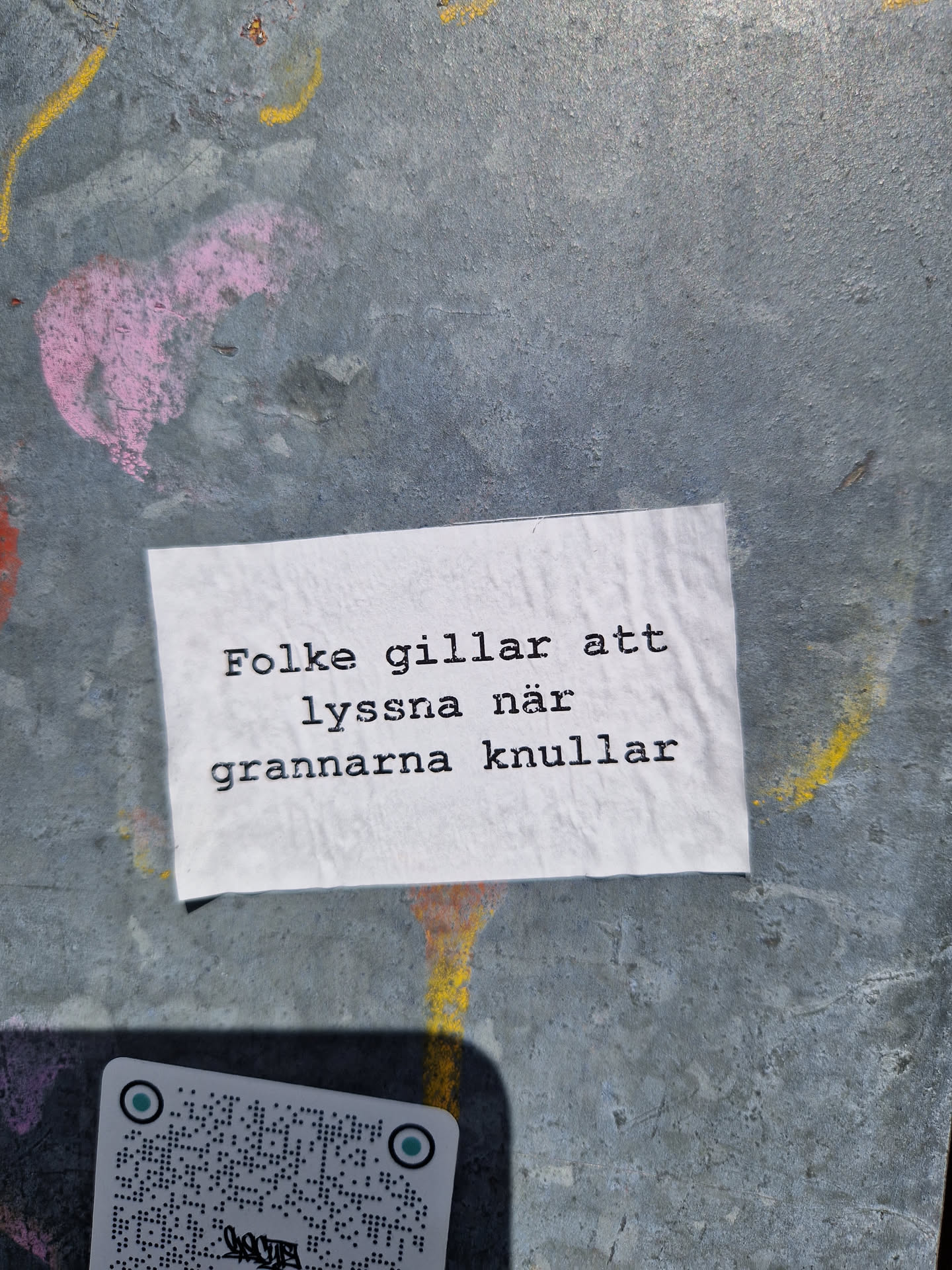 A white sticker on a gray wall with text in Swedish.  The text translates to: "People like to listen when the neighbors fuck." A QR code is visible below the sticker.