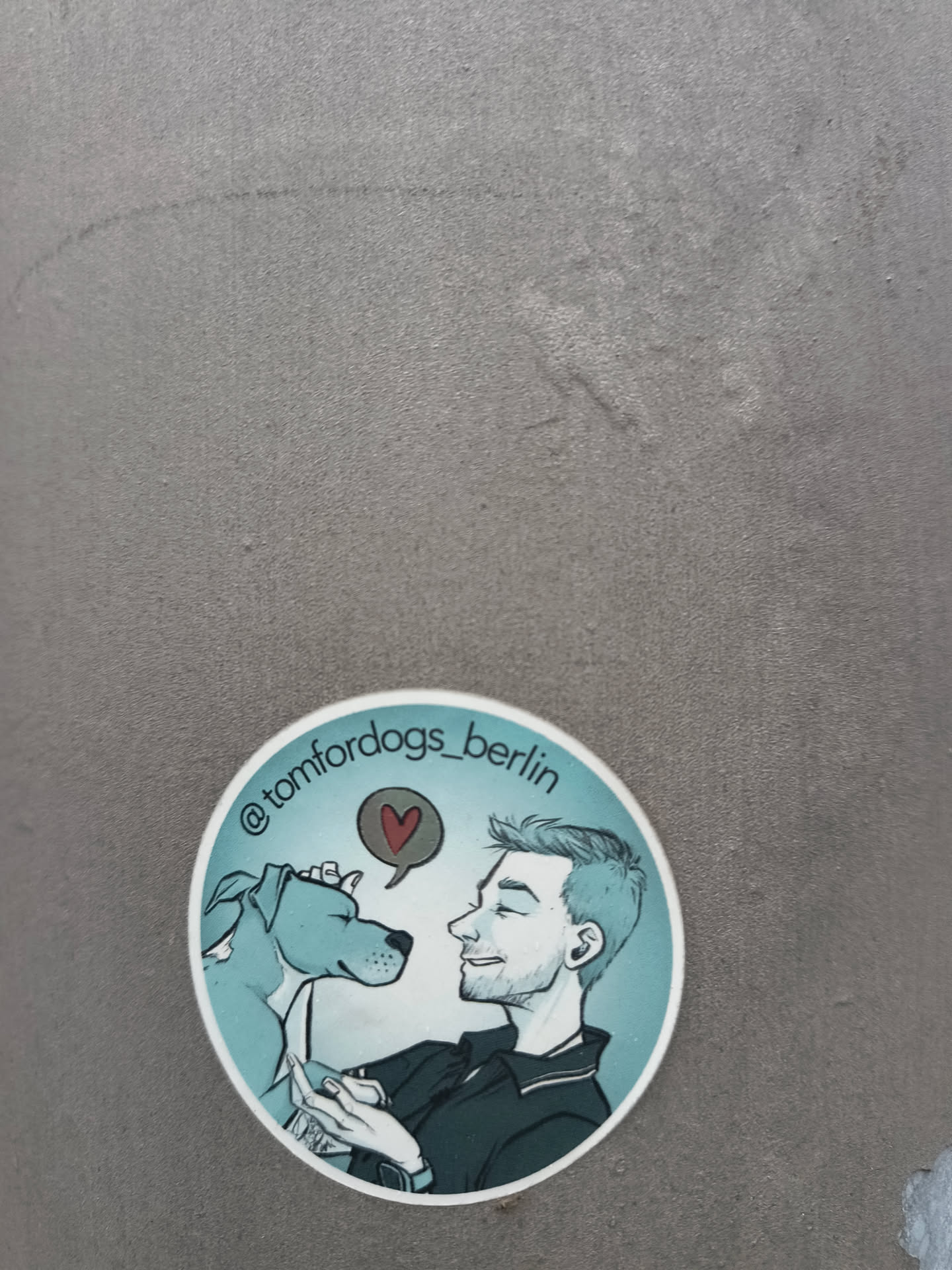 The sticker is circular with a white border. It features a stylized illustration of a man and a dog. The man is on the right, depicted in profile with his eyes closed and a smile, wearing a dark collared shirt. The dog, on the left, is a light blue pitbull-type breed, also in profile, with its eyes closed and a red heart symbol in a speech bubble above its head. The background is a gradient of light blue. The text '@tomfordogs_berlin' is written in a curve along the top edge of the sticker.
