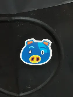 The sticker features a stylized, cartoon-like pig's head with a predominantly vibrant blue color. It has a rounded, irregular shape with small, orange-tipped ears at the top. The face includes a prominent yellow snout with two dark brown oval nostrils. Its eyes are white ovals with small black pupils, one slightly larger than the other, giving it a playful or slightly surprised expression. A lighter blue patch is visible on the right side of its head, and simple blue curved lines serve as eyebrows. The entire design is outlined with a clean white border, making it stand out against the dark background.