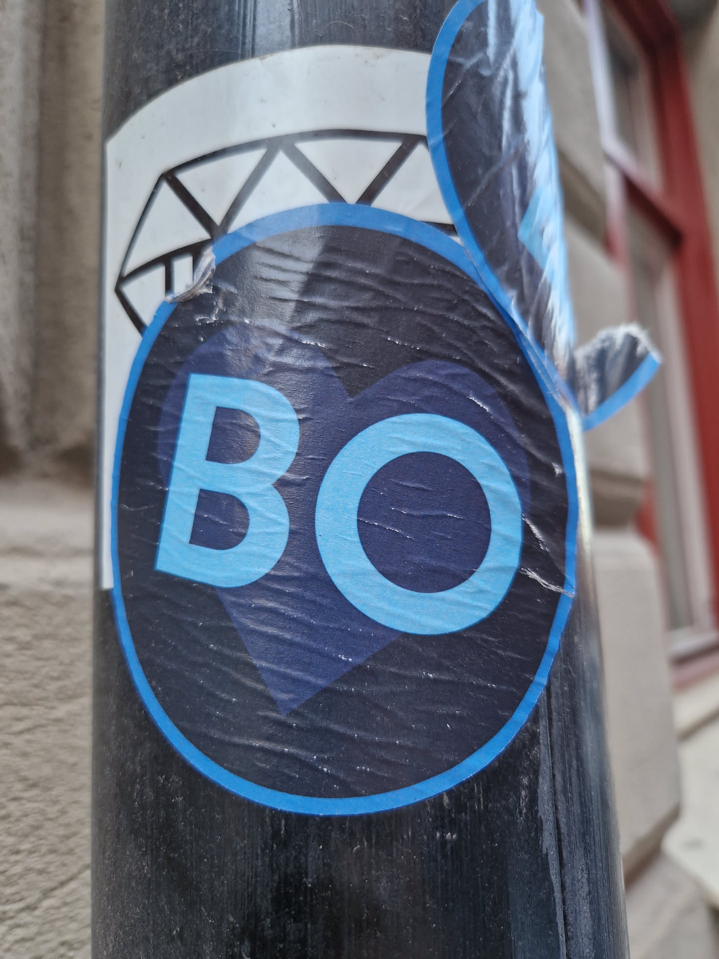 The sticker is primarily dark blue, shaped like a stylized heart or an inverted teardrop, outlined with a thinner light blue border. Centered within the dark blue area are the capital letters "BO" in a light blue, sans-serif font. The 'O' is slightly larger than the 'B' and partially overlaps it. The sticker shows visible signs of wear, including wrinkles and slight peeling at the edges, suggesting it has been exposed to the elements. It is affixed to a dark, possibly metallic, pole. Another partial white sticker with black geometric patterns is visible behind and above the main sticker.