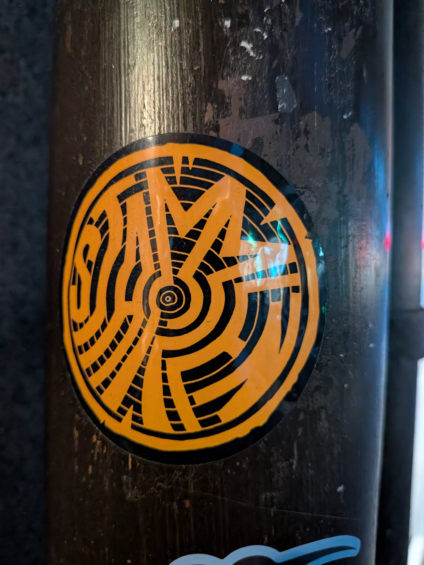 The sticker is circular with a black border. The interior is predominantly bright orange with bold black lines forming an abstract, geometric pattern. The design features concentric circles and radiating lines that create a sense of depth and complexity. The overall aesthetic is graphic and eye-catching.