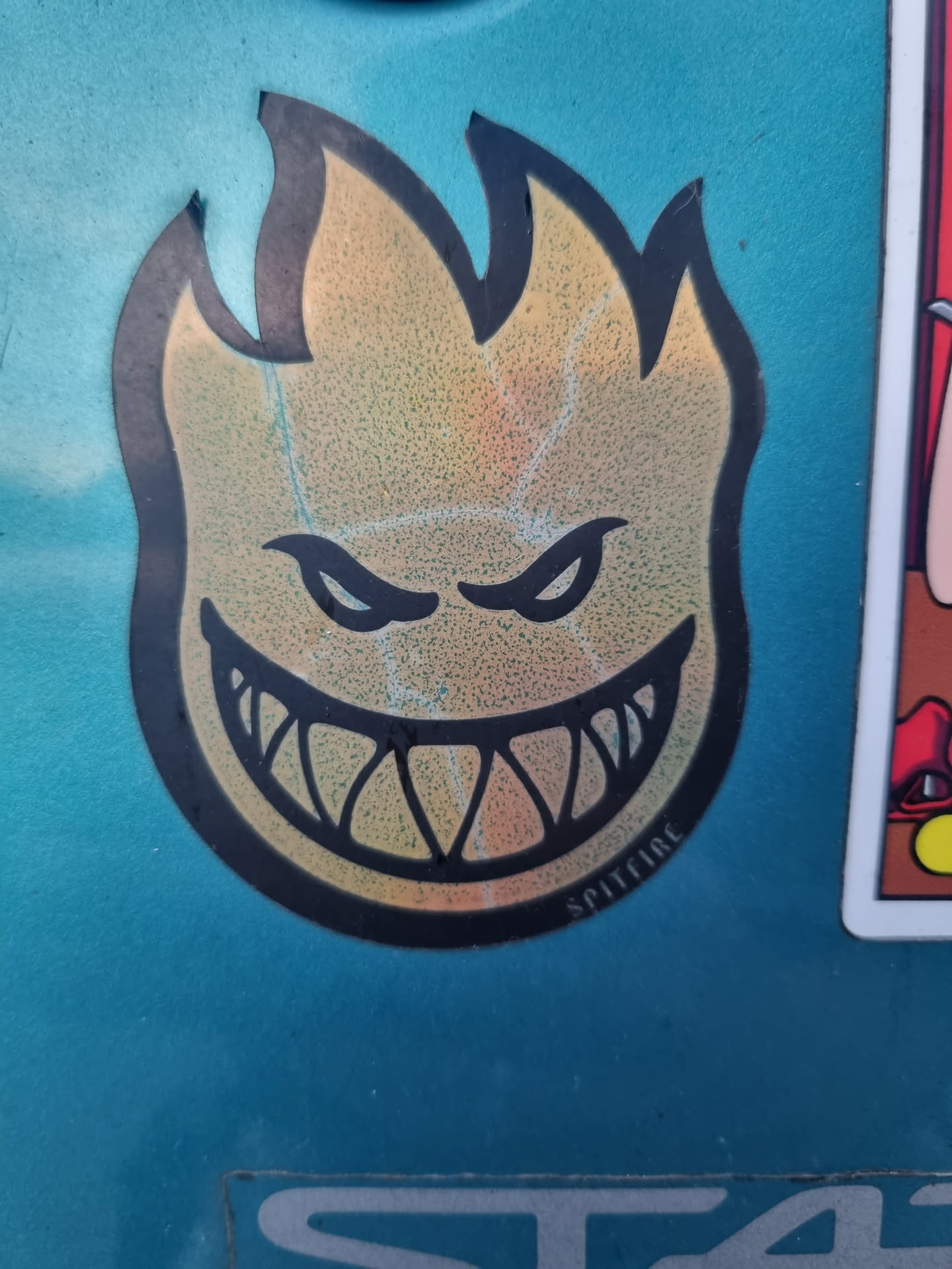 A classic Spitfire Wheels sticker featuring their iconic flame head logo. The sticker has a holographic or iridescent effect, giving it a shiny, multicolored look.  The flame head has sharp teeth and angry eyes.