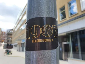 A black and gold sticker featuring the Helsingborgs IF logo and the year 1907. The sticker is affixed to a metal pole on a city street.