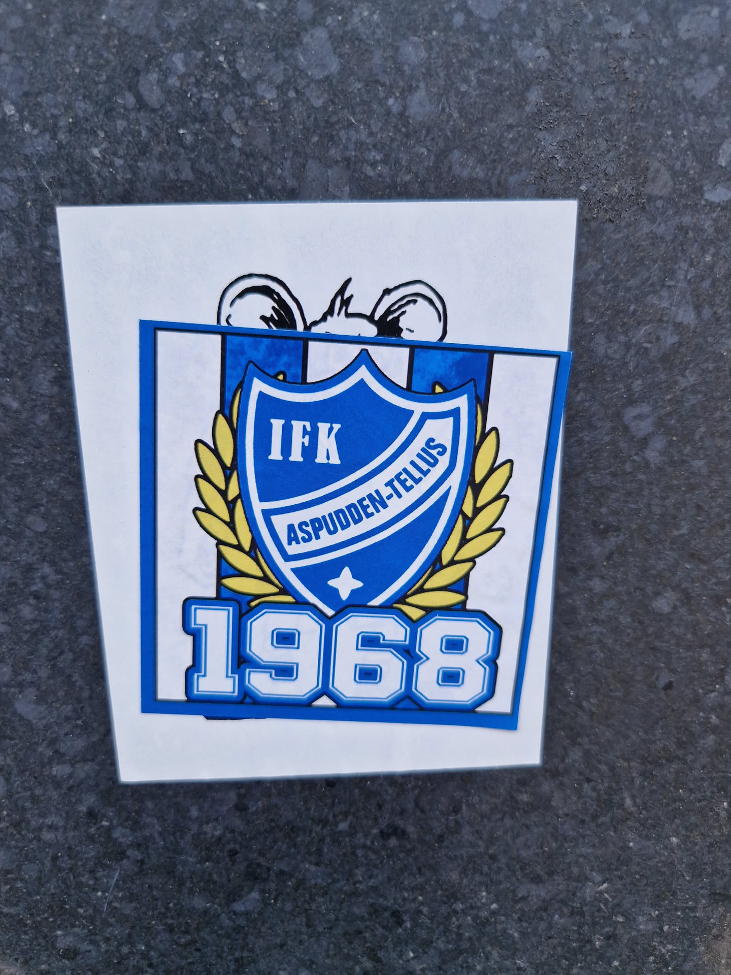 A blue and gold sticker featuring the logo of IFK Aspudden-Tellus, a Swedish sports club founded in 1968. The logo includes a shield with the club's initials and name, surrounded by a laurel wreath.  The year 1968 is prominently displayed below the logo. A cartoon mouse's ears are visible at the top of the sticker.