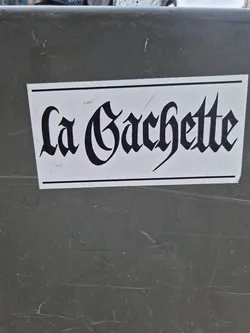 A sticker with the text "La Gachette" written in a black, old-style font on a white background. The sticker is affixed to a dark gray surface.
