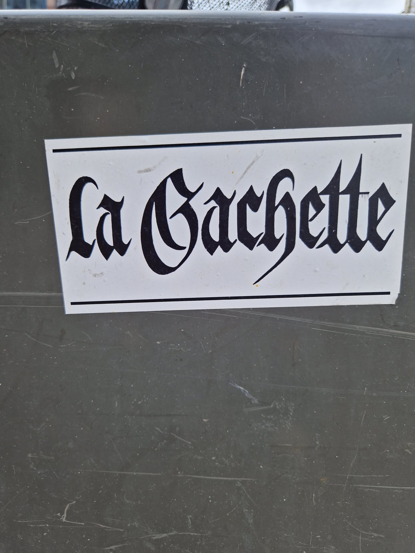 A sticker with the text "La Gachette" written in a black, old-style font on a white background. The sticker is affixed to a dark gray surface.