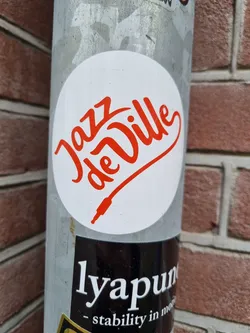 Round white sticker with red text saying "Jazz de Ville" in a stylized script font.  A small red audio jack plug is also part of the design. 