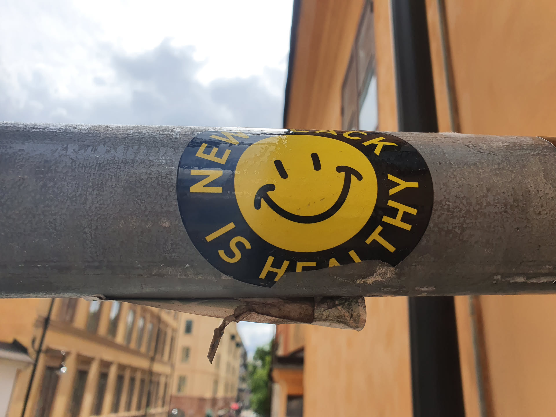 A round black and yellow sticker with a smiley face and the text "NEW IS HEALTHY" stuck on a gray metal pole.