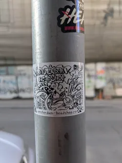A black and white sticker with a doodle-like drawing and the text "Amsterdam Headshop" and "amsterdam-headshop.ro". The drawing includes elements like a mushroom cloud, various monsters, buildings, and smoking paraphernalia.