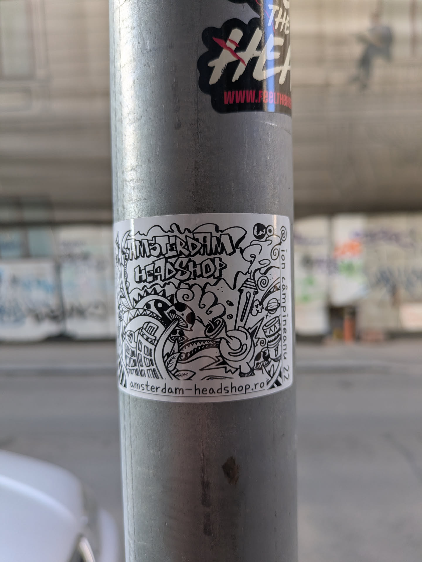 A black and white sticker with a doodle-like drawing and the text "Amsterdam Headshop" and "amsterdam-headshop.ro". The drawing includes elements like a mushroom cloud, various monsters, buildings, and smoking paraphernalia.