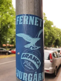 A blue and white Fernet Branca sticker featuring the brand's iconic eagle and globe logo. The sticker is affixed to a metal pole, and part of the text 'Jurgarn' is visible at the bottom. This vintage design is a nod to the classic branding of Fernet Branca, an Italian bitter.
