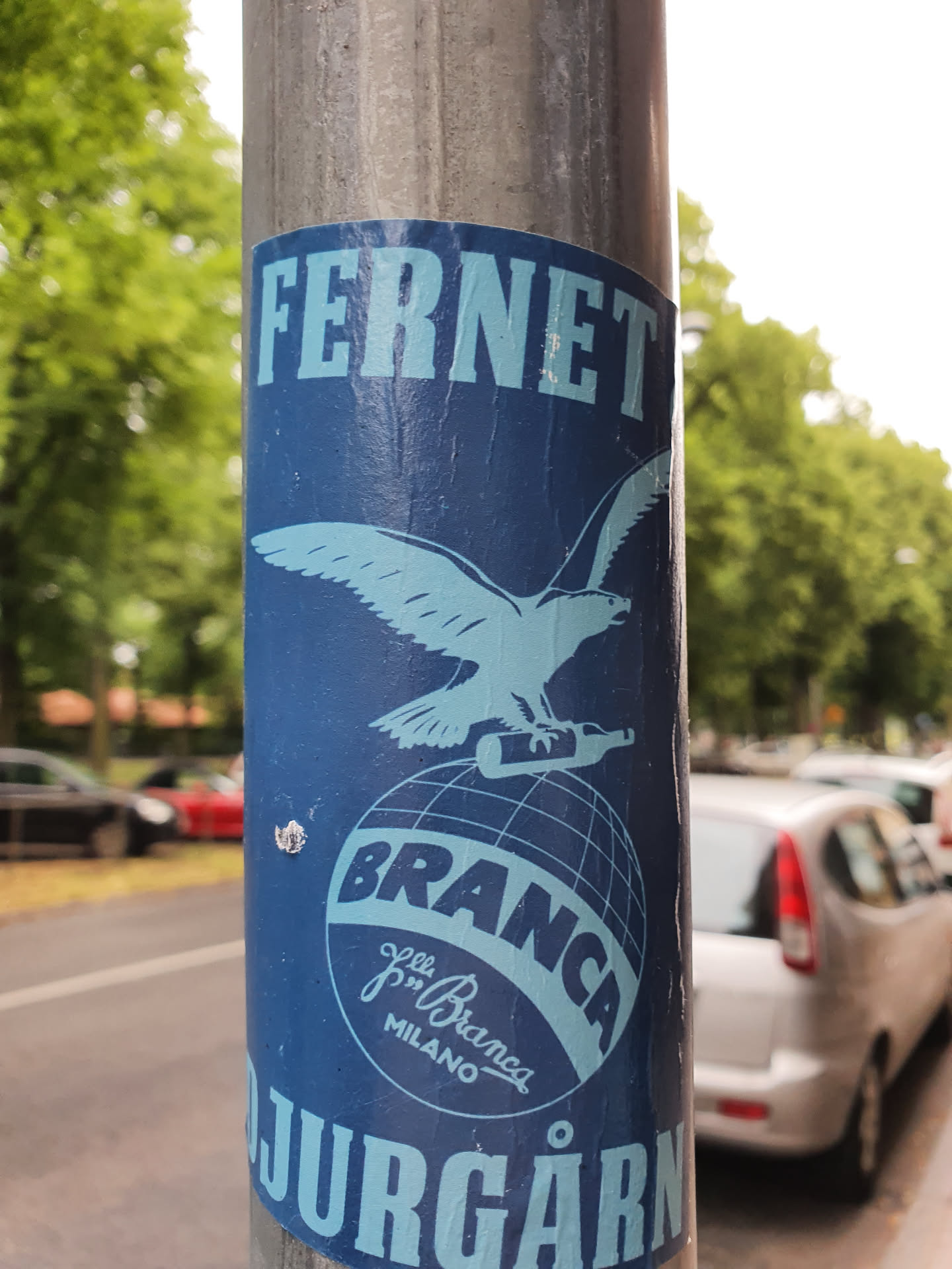 A blue and white Fernet Branca sticker featuring the brand's iconic eagle and globe logo. The sticker is affixed to a metal pole, and part of the text 'Jurgarn' is visible at the bottom. This vintage design is a nod to the classic branding of Fernet Branca, an Italian bitter.