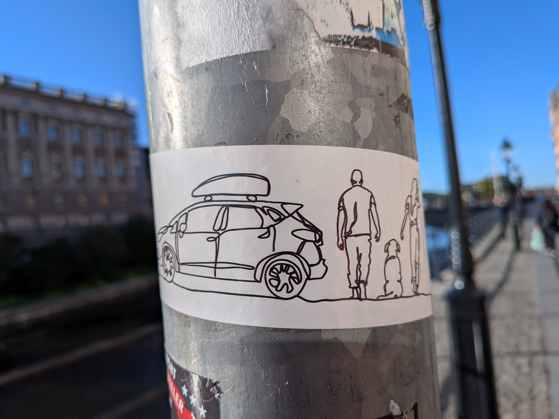 The sticker is a minimalist, black and white outline drawing on a white rectangular background. It depicts a car with a roof box, followed by a silhouette of a person, a dog sitting, and another person standing. The style is simple and linear, with no shading or color fill.