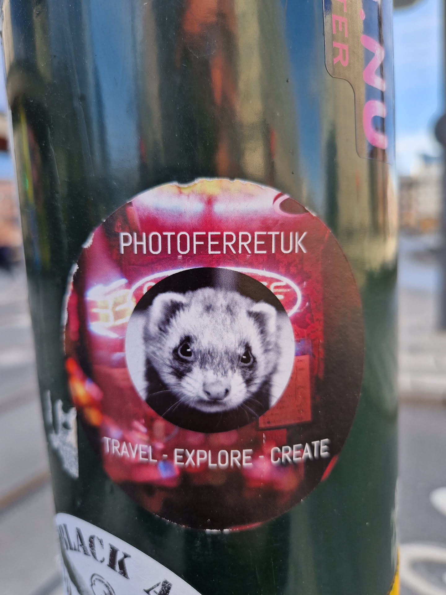 A round sticker featuring a ferret's face against a blurry background of neon lights.  The text "PHOTOFERRETUK" is at the top, and "TRAVEL - EXPLORE - CREATE" is at the bottom. 