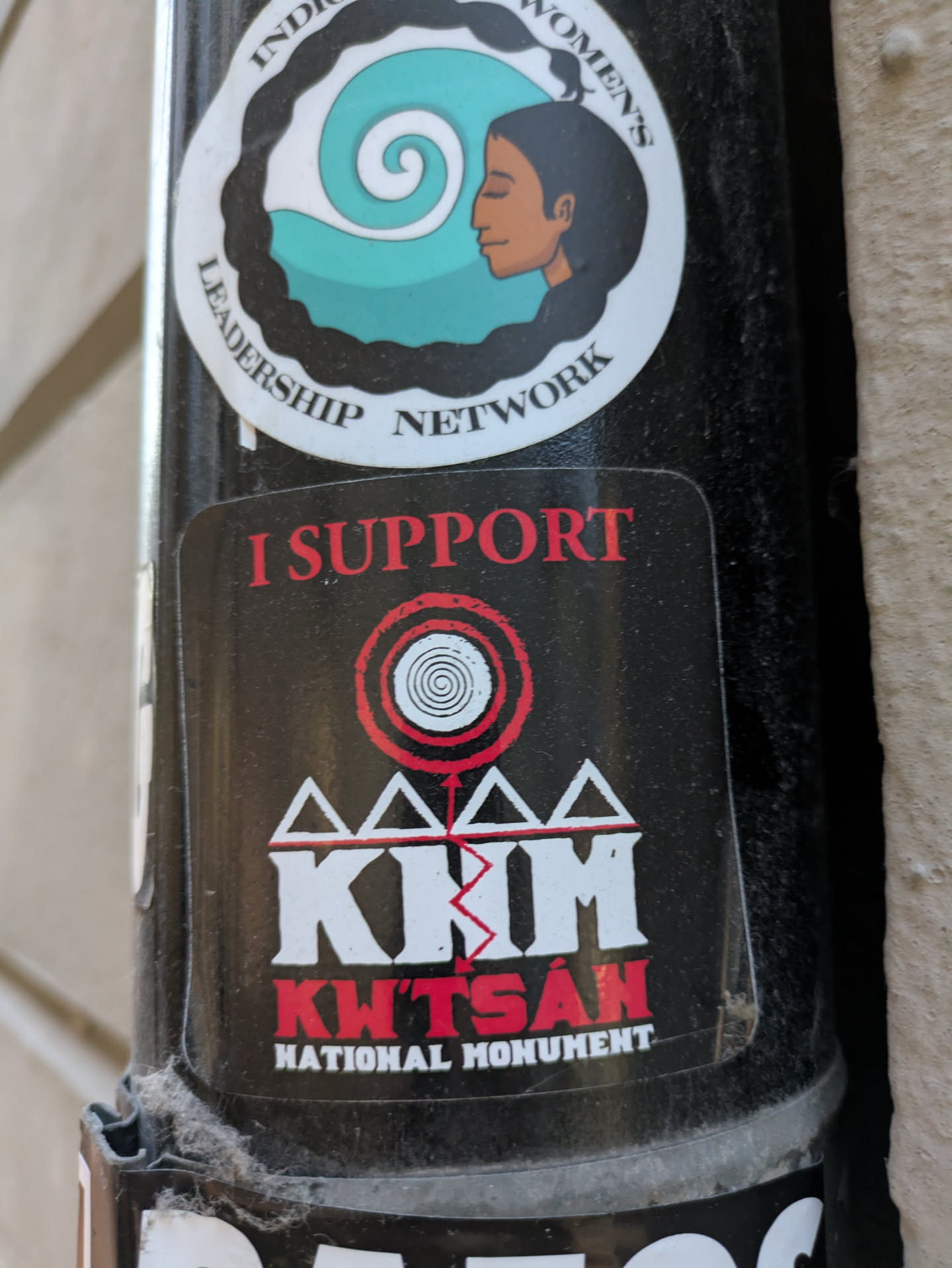 The sticker is black with red and white text and graphics. At the top, it reads 'I SUPPORT' in red capital letters. Below this is a graphic with concentric red and white circles, resembling a target or a stylized vortex. Underneath this symbol are four white triangles, connected at their bases, forming a stylized mountain or pyramid shape. The letters 'KWT'SAN' are prominently displayed in white, bold, block letters with a jagged red line cutting through the 'M'. Below this, in smaller red capital letters, it says 'NATIONAL MONUMENT'. The sticker appears to be slightly worn and has some dust or debris on its surface.
