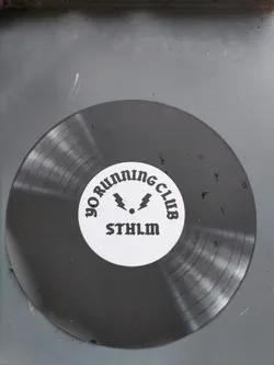 A dark gray vinyl sticker in the shape of a record. It has a white circle in the center with the text "YO RUNNING CLUB" around a symbol and the abbreviation "STHLM".