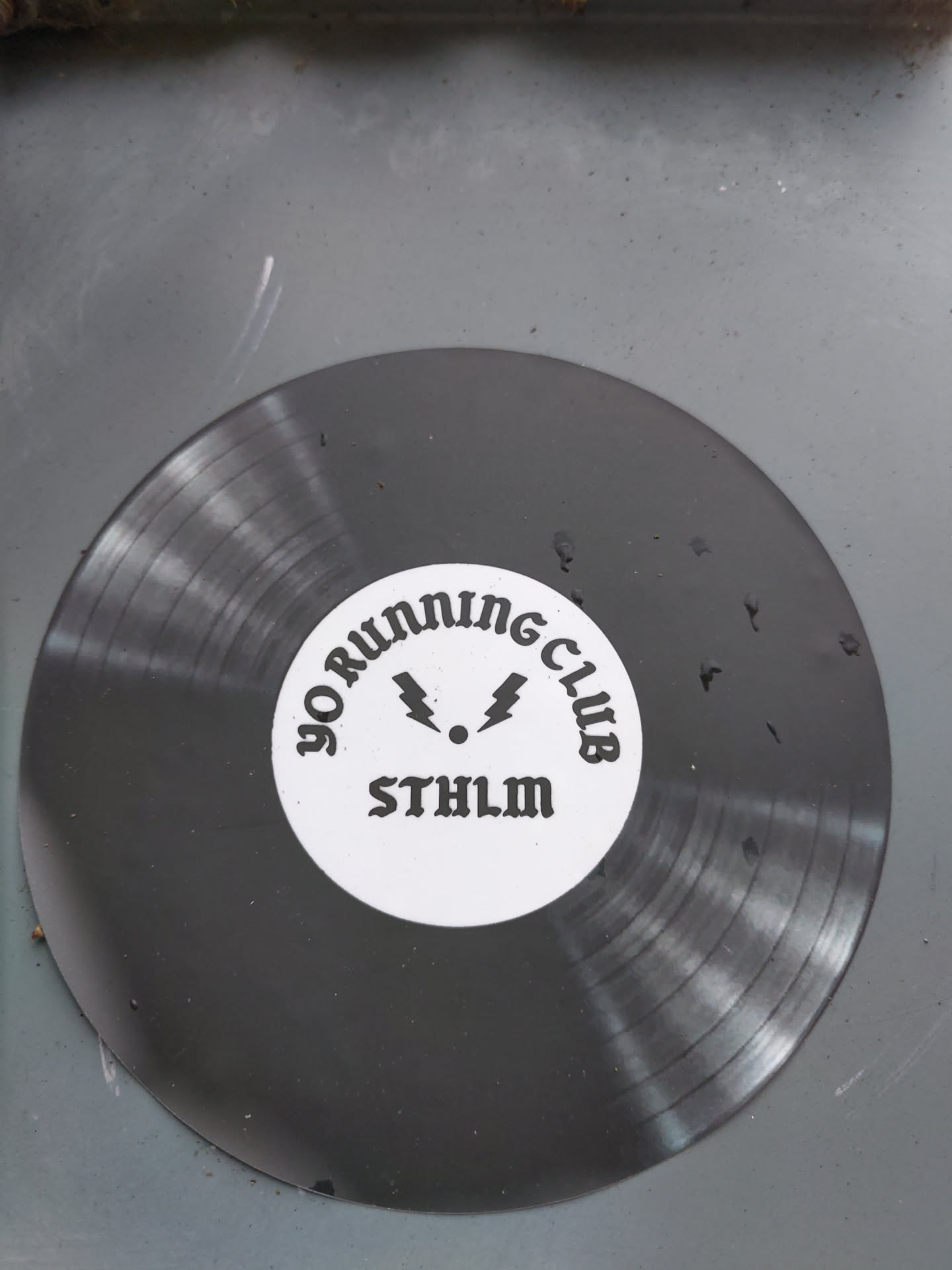 A dark gray vinyl sticker in the shape of a record. It has a white circle in the center with the text "YO RUNNING CLUB" around a symbol and the abbreviation "STHLM".