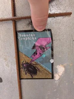 A rectangular sticker with a black border featuring a drawing of a black bug at the bottom and a pink lightning bolt with a skull in the middle. The background is blue with the words "Nomadak Graphiks" on the top left and "Serigrafia Brutal" on the bottom right.