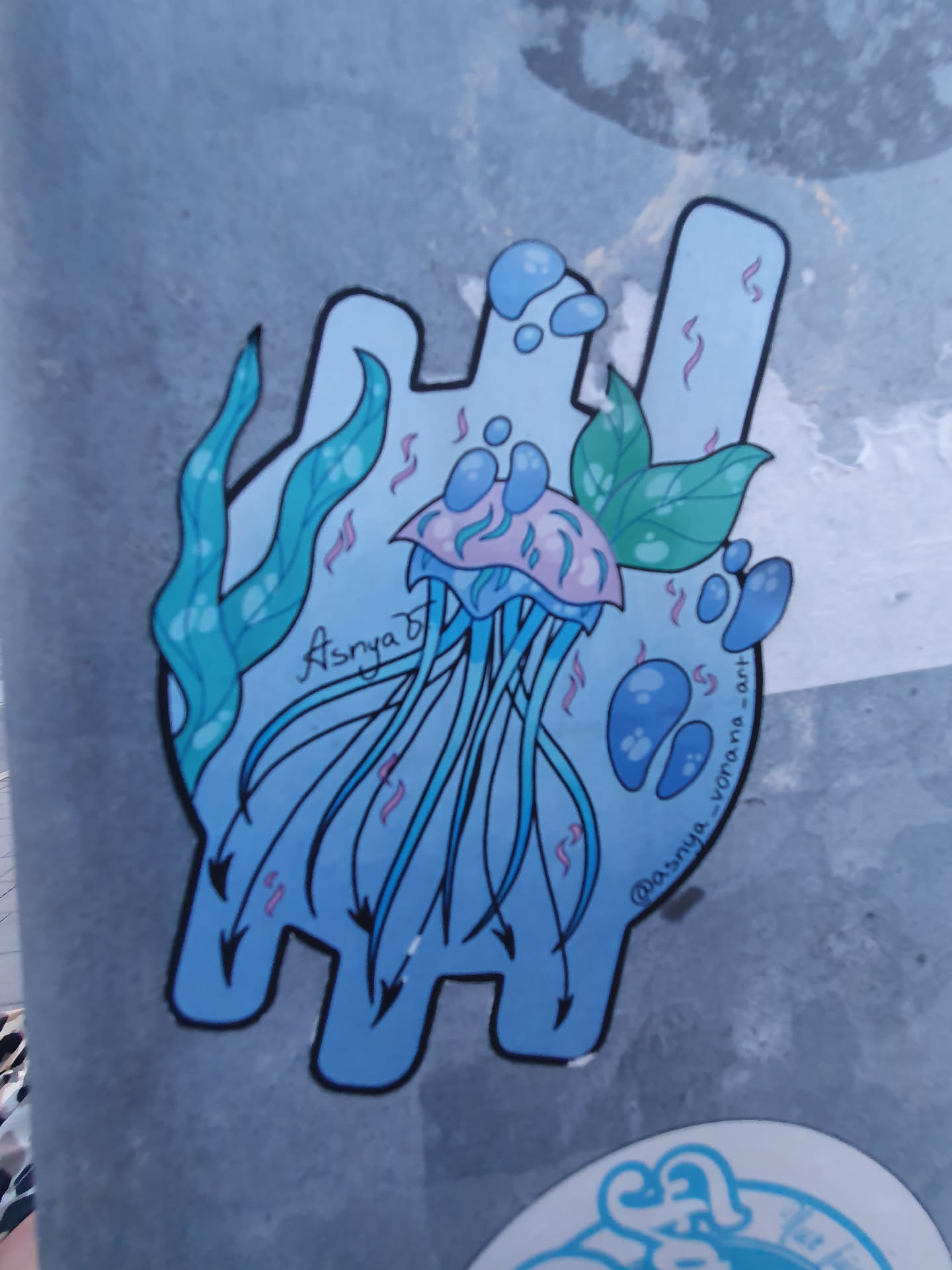 A cute jellyfish sticker with seaweed and bubbles in light blue and pink colors. The sticker is in a unique shape, and has the artist's name, Asrya, written on it. There is also an instagram handle @asrya_vorona_art on the sticker.