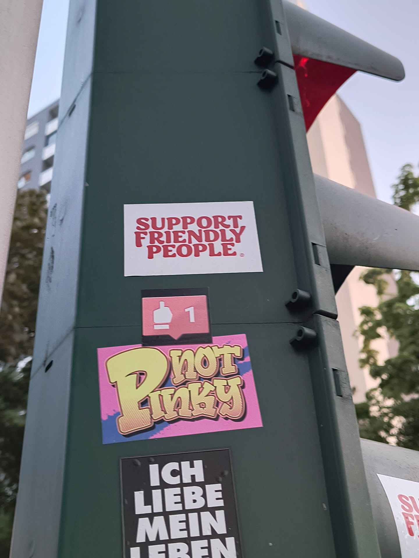 The sticker is composed of three distinct elements layered on a dark green pole. The topmost element is a white rectangular sticker with bold red text that reads 'SUPPORT FRIENDLY PEOPLE.' Below this, a smaller sticker displays a pink speech bubble icon with a white thumbs-up symbol and the number '1' inside, resembling a social media 'like' notification. The largest and most vibrant sticker is situated at the bottom, featuring the word 'Pinky' in a graffiti-style font. The letters are yellow with a textured gradient, outlined in black and white, and set against a bright pink background with blue and purple abstract shapes. Another black sticker with white text is partially visible below the 'Pinky' sticker.