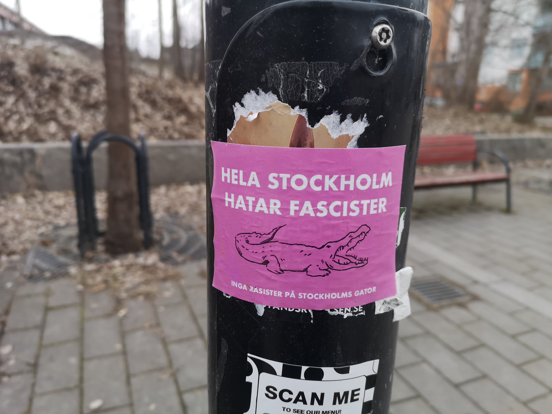 A vibrant pink sticker adhered to a black pole. The sticker features bold white uppercase text at the top, reading 'HELA STOCKHOLM HATAR FASCISTER'. Below this, a black outline illustration of a cartoonish alligator or crocodile with its mouth open is prominently displayed. Further below the alligator, smaller white text reads 'INGA RASISTER PÅ STOCKHOLMS GATOR'. The sticker shows signs of wear, with some peeling at the top edge, revealing a lighter brown paper underneath.