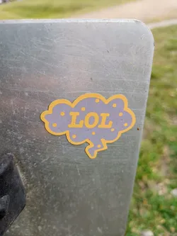 A small sticker of a purple cloud with the letters LOL in yellow. The sticker is on a scratched metal surface.